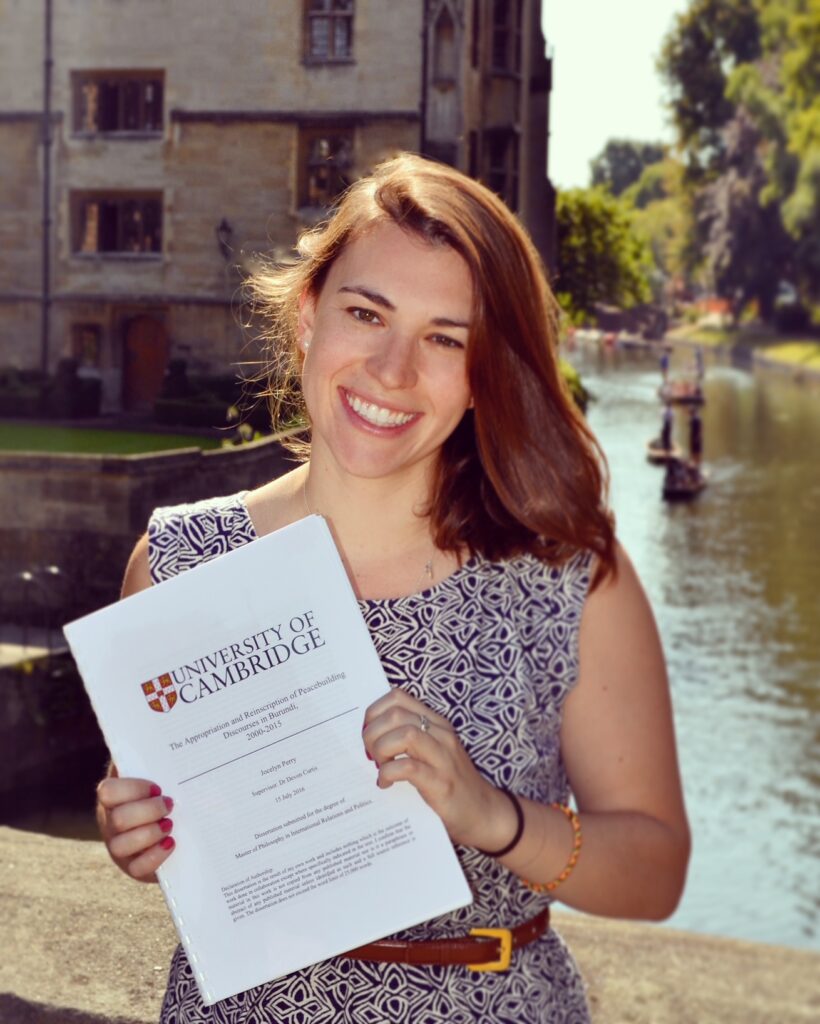 Jocelyn holding her MPhil dissertation with the Cambridge logo on the front cover standing in front of the River Cam with a punt boat behind her