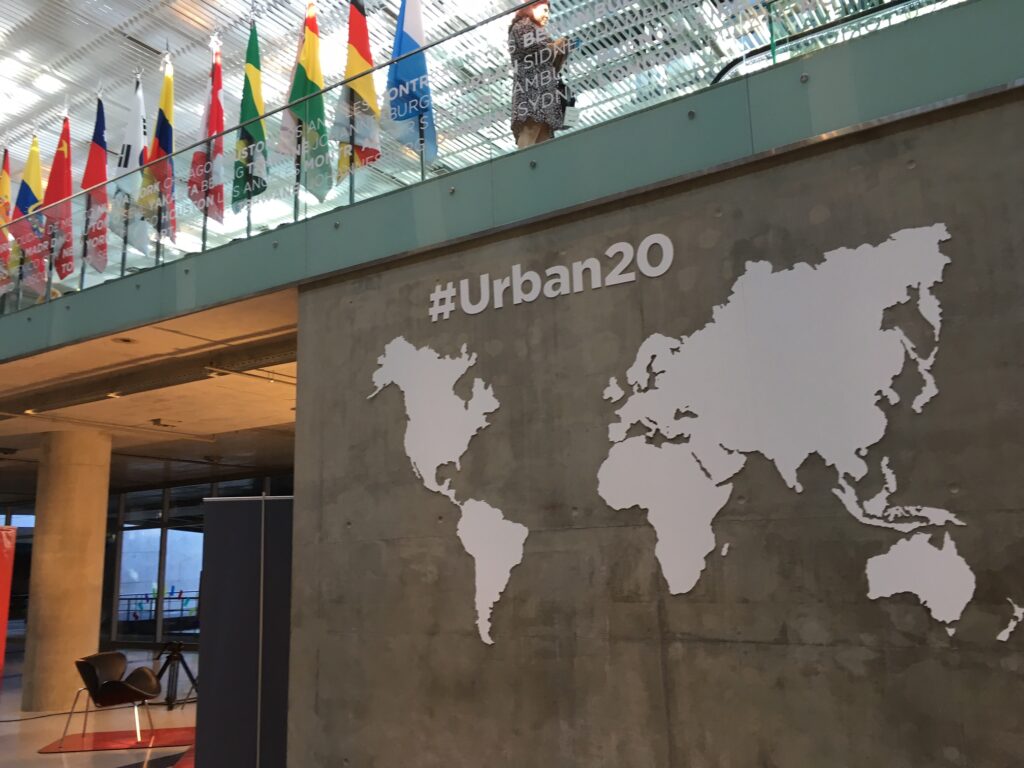 White sign saying #Urban20 with cutout map of world on concrete wall with flags in background on upper level in light-filled convention center in Buenos Aires, Argentina