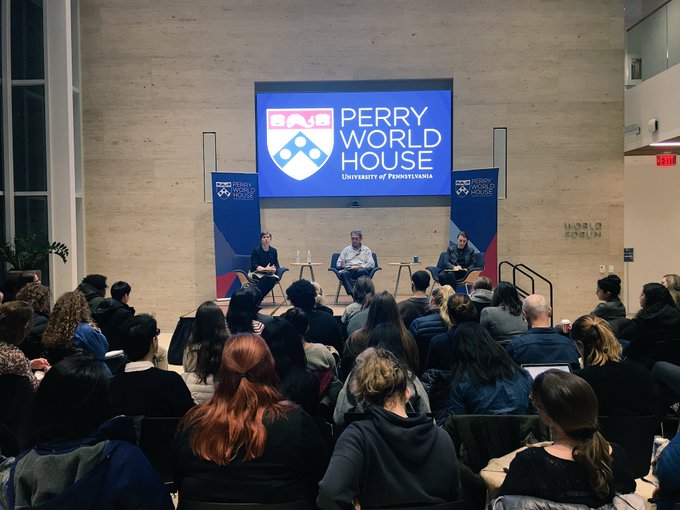 Three speakers on a stage with an audience in the foreground, with two banners and a lit-up screen saying Perry World House against a marble wall in background.